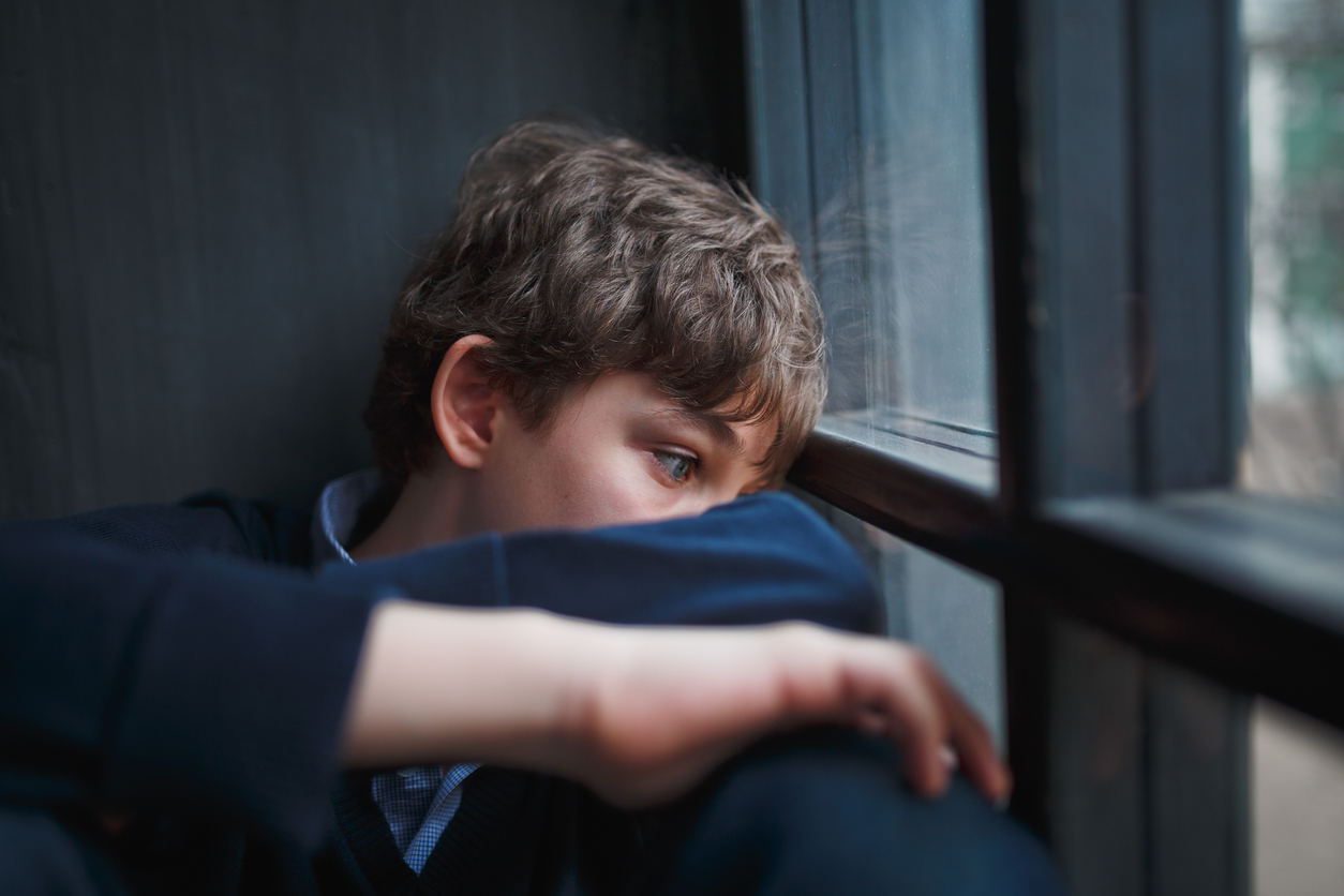 Pensive sad boy teenager in a blue shirt and jeans sitting at the window and closes his face with his hands. barn-unge-mistrives-omsorgs-svigt-snak-om-det-psykoterapi-vivi-hinrichs-aarhus
